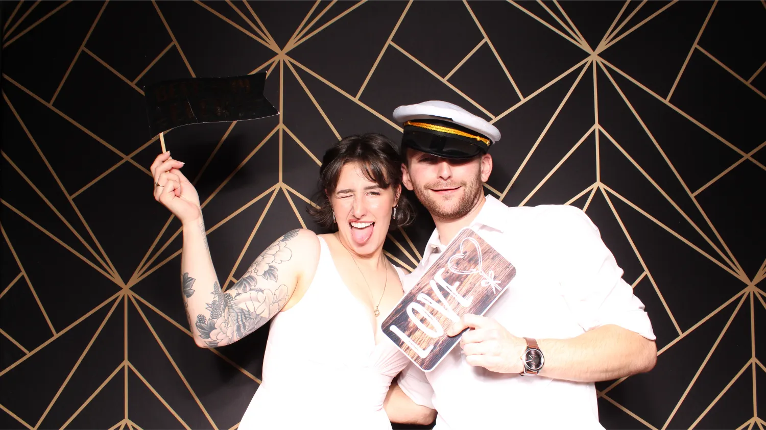 A smiling couple in a photo booth, the woman winking and sticking out her tongue while holding a black sign, and the man wearing a captain's hat holding a sign that says LOVE.