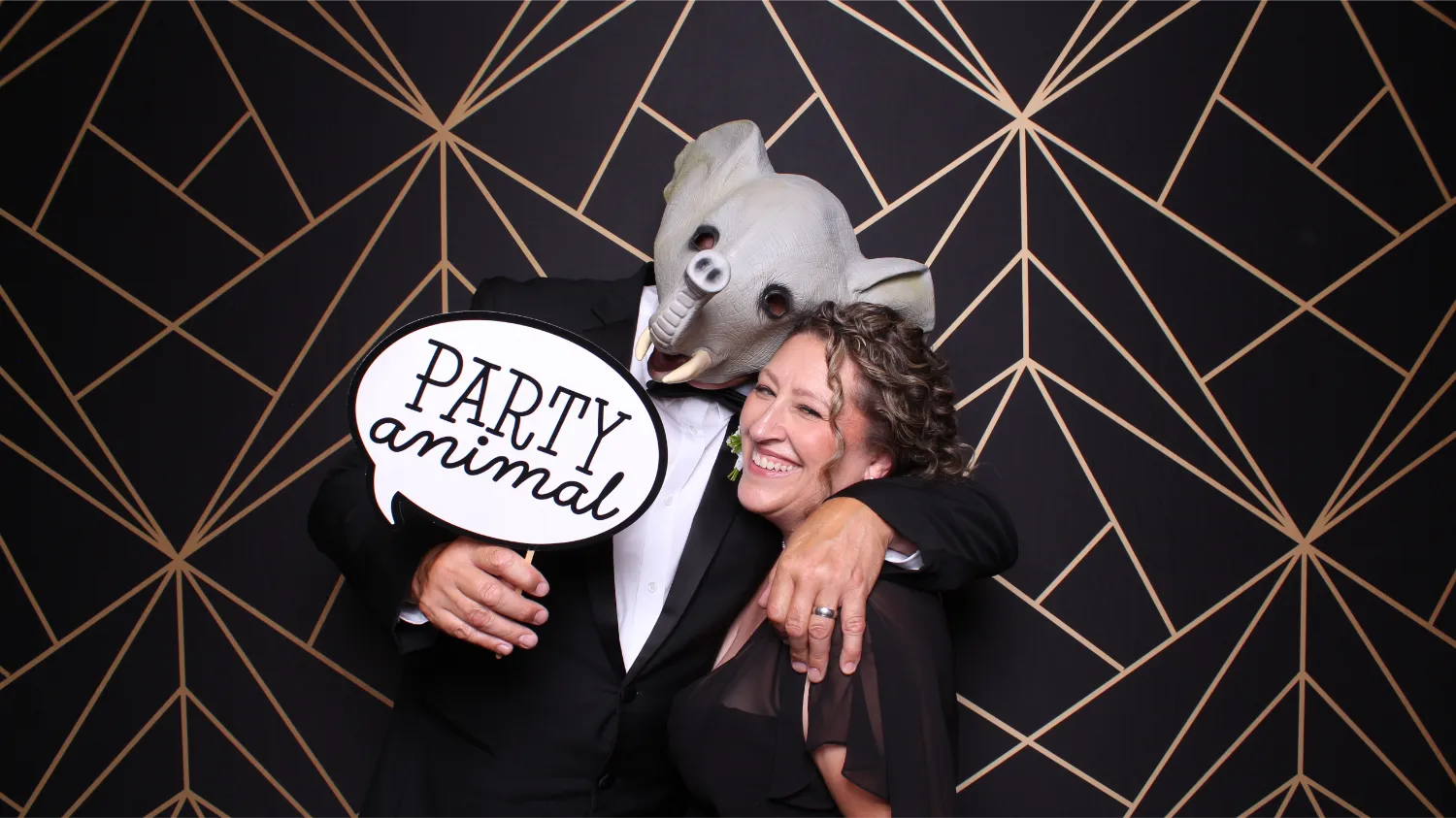 Person in an elephant mask in a tuxedo embraces a smiling woman in a black dress holding a sign that says 'PARTY animal' against a geometric black and gold background.