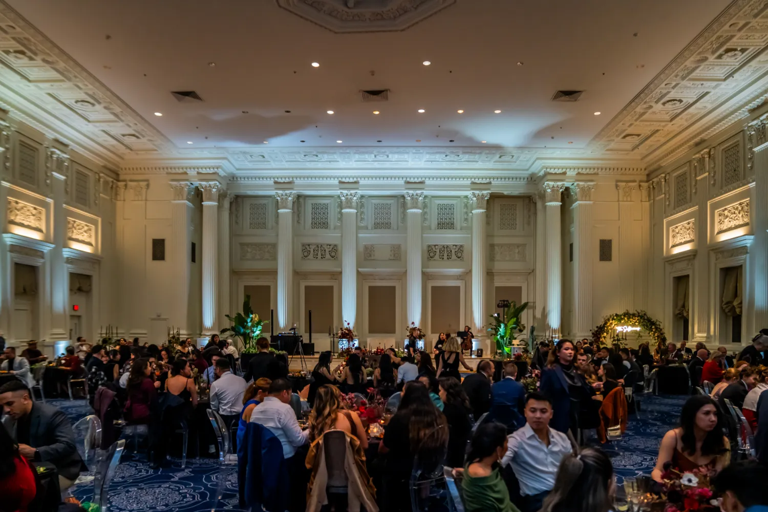 Large elegant banquet hall with white pillars and ornate ceiling filled with seated guests at round tables dressed for a formal event.