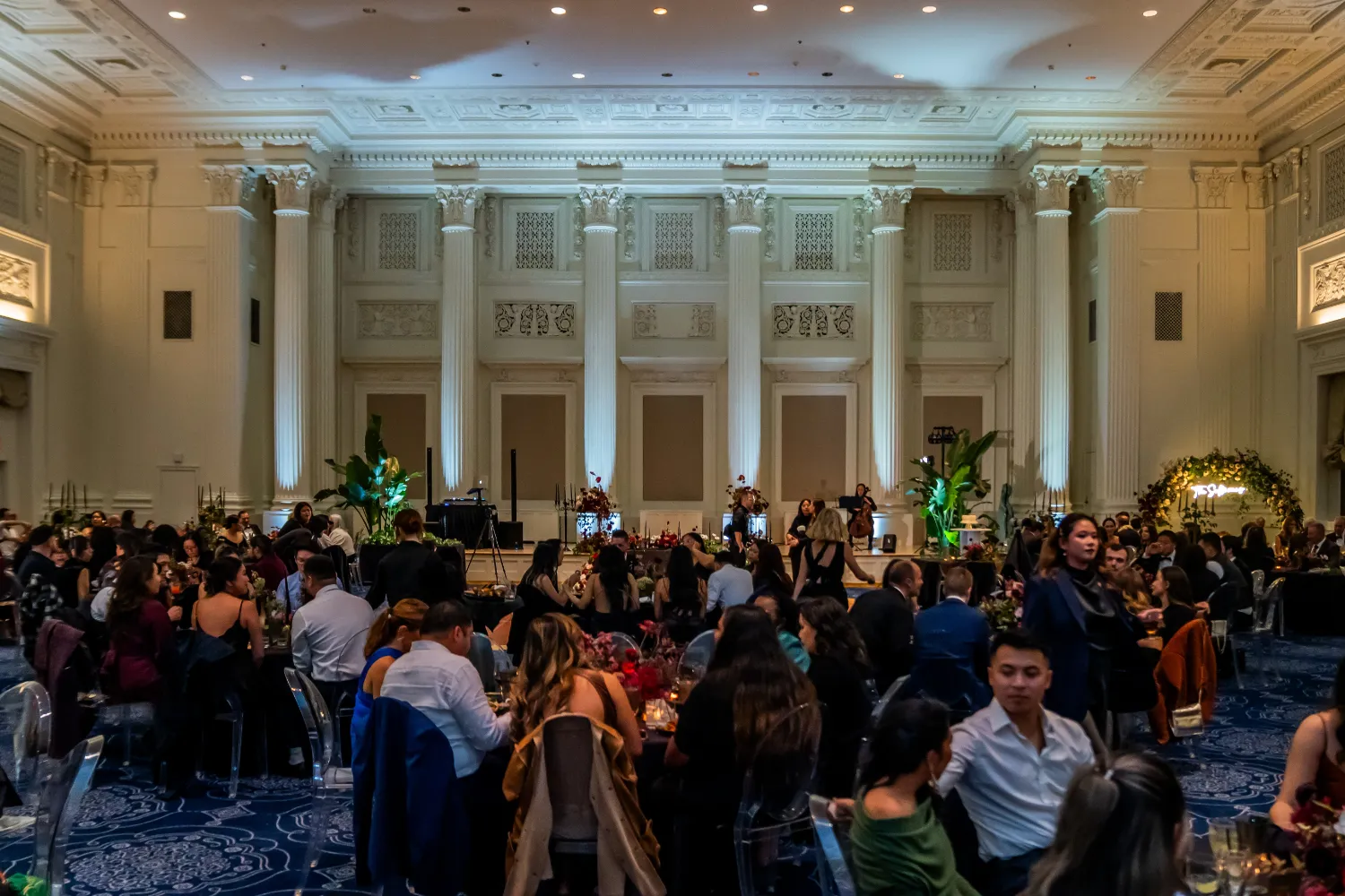 Large elegant banquet hall with tall white columns and detailed ceiling, filled with guests seated at round tables during an event.