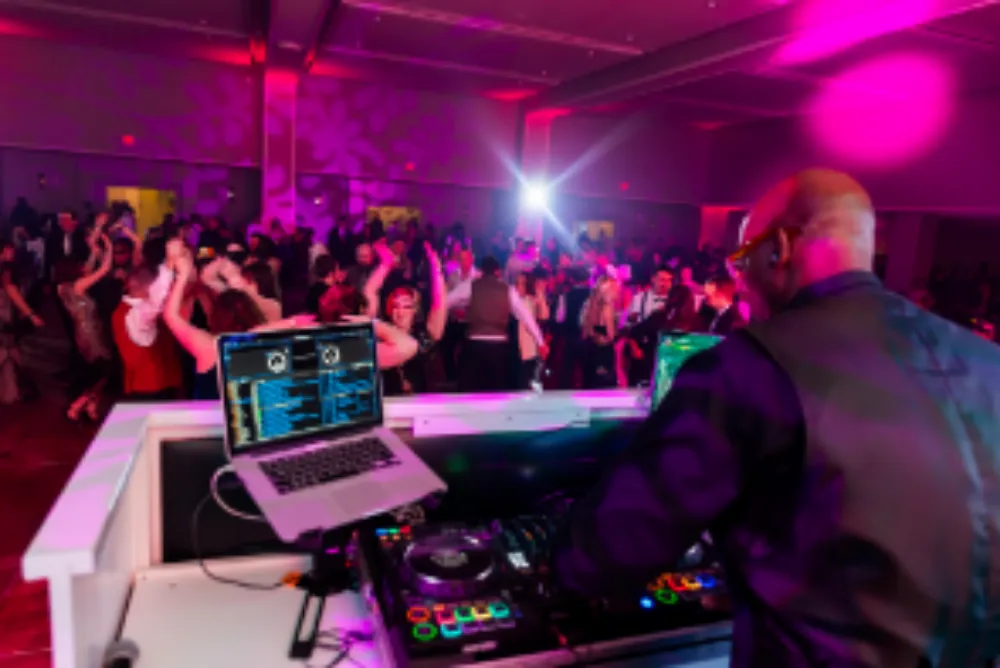 DJ performing at a corporate event with a laptop and mixer, colorful lights and a crowd dancing in the background.