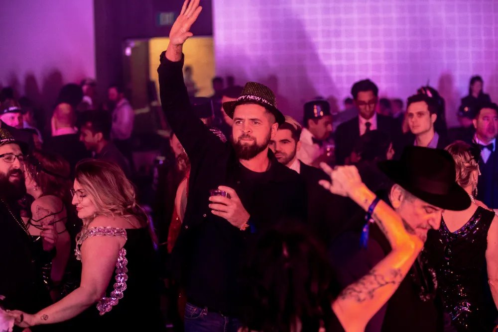 Man wearing a 'Happy New Year' hat raising his hand while holding a drink at a crowded party with people dancing.