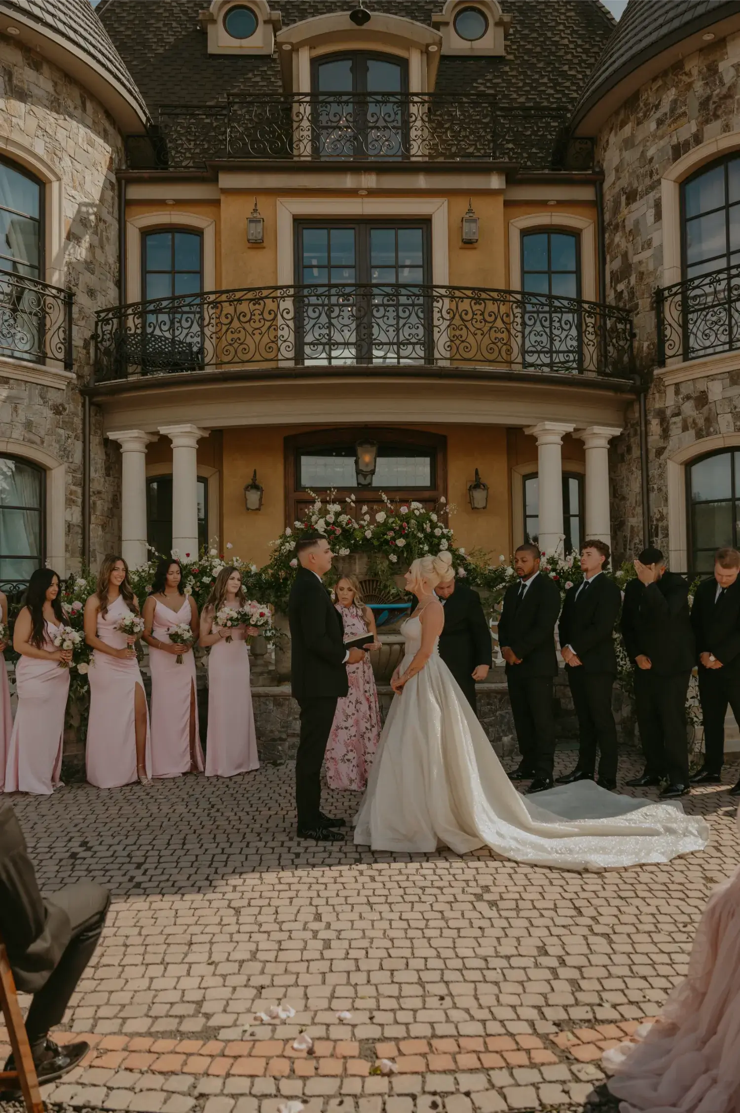 Bride and groom standing at the altar during an outdoor wedding ceremony in front of a stone mansion, surrounded by bridesmaids in pink dresses and groomsmen in black suits.