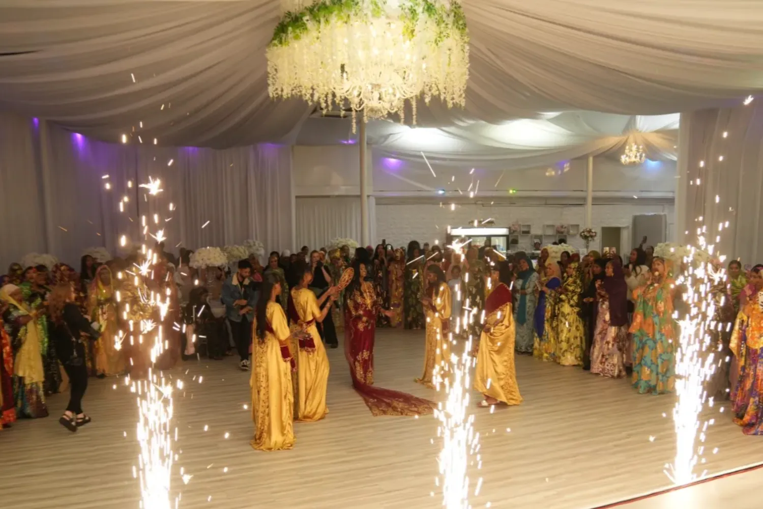 Indoor Indian wedding celebration with women in traditional attire dancing surrounded by bright sparklers and a chandelier overhead.