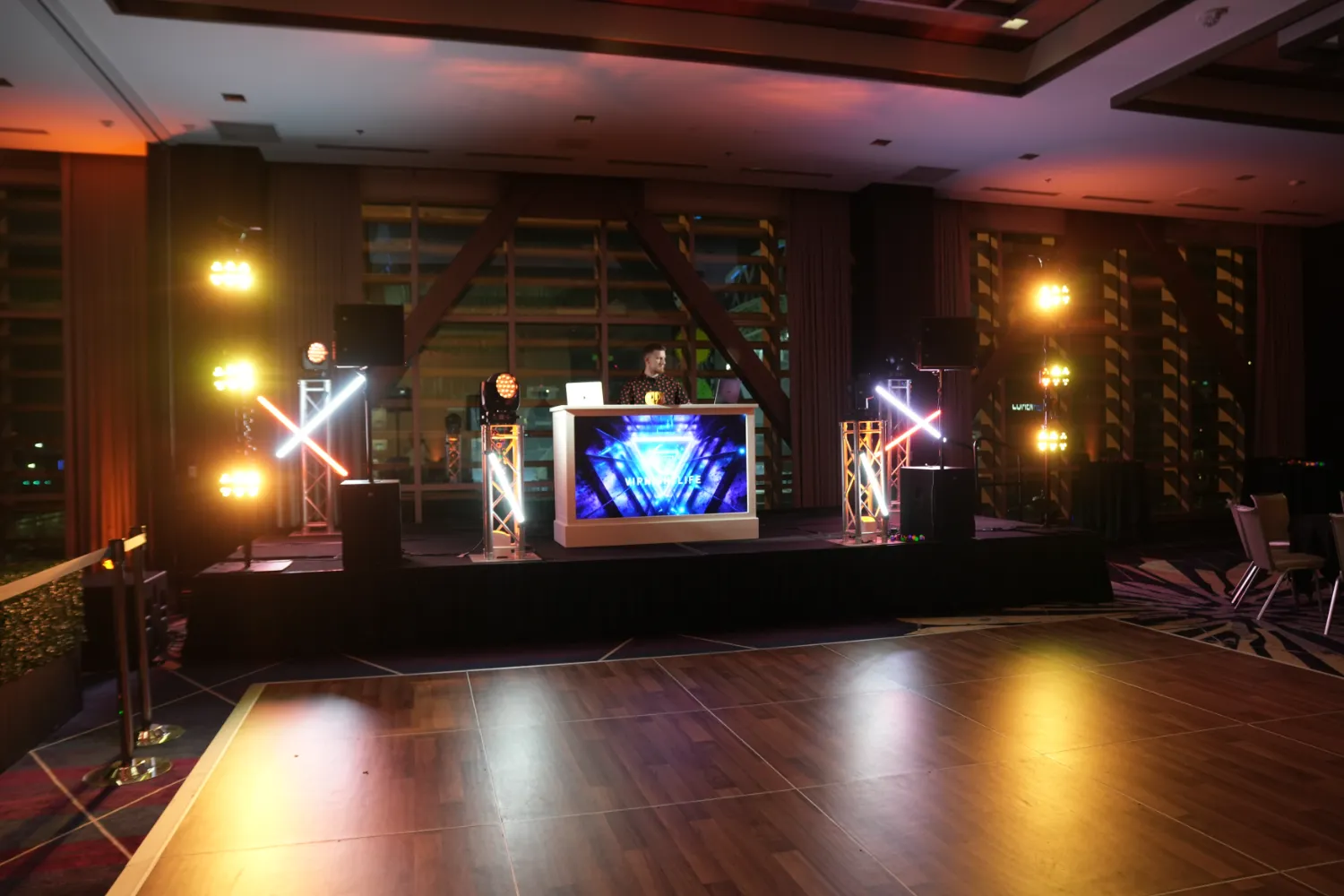 DJ standing behind illuminated booth on stage with colorful lighting in an event space with wooden dance floor.