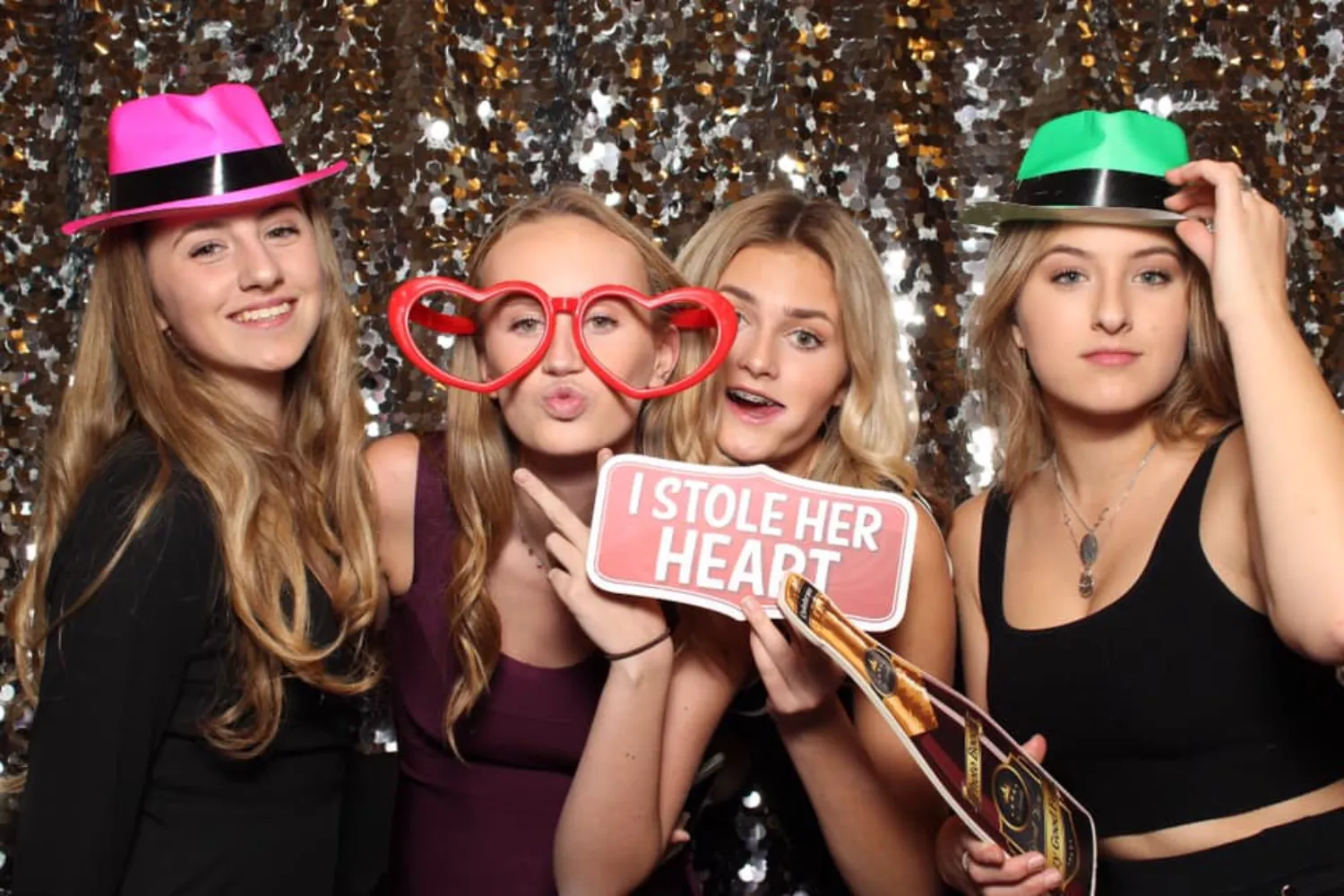 Four teenage girls posing in a photo booth with festive hats and props, one holding a sign reading 'I STOLE HER HEART.'