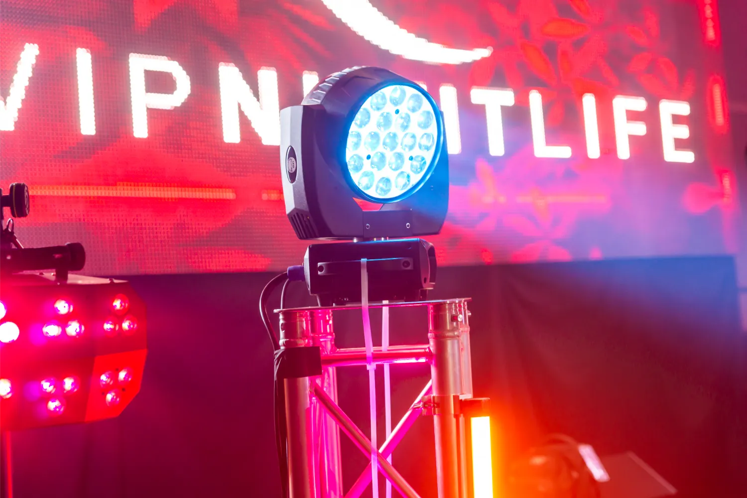 Stage lighting setup with a blue LED moving head light on metal truss and red neon lights, with a blurred lit sign reading VIP NIGHTLIFE in the background.