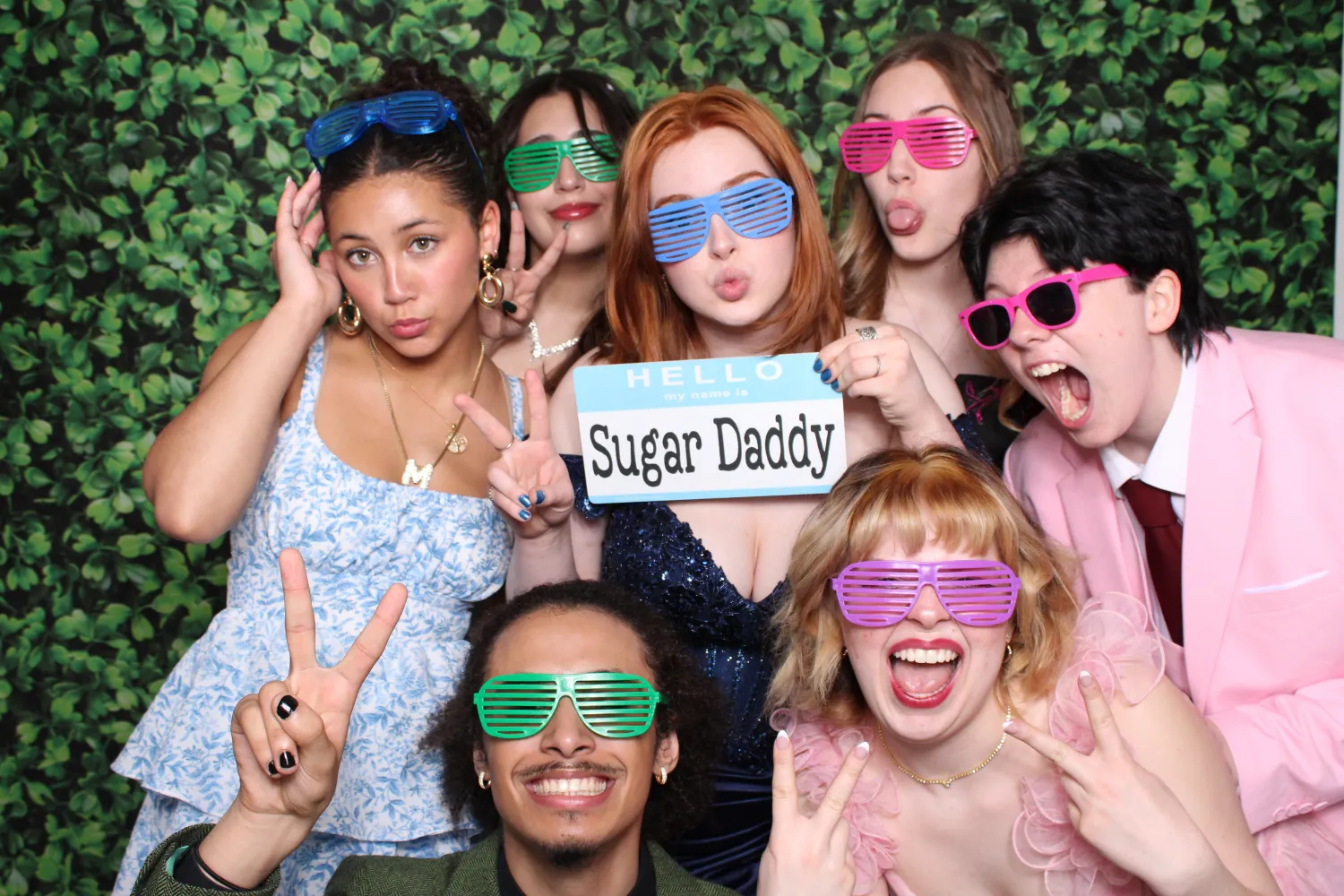 Group of seven young people at a photo booth wearing colorful shutter shades, making peace signs and playful faces, one holding a 'Hello my name is Sugar Daddy' sign.