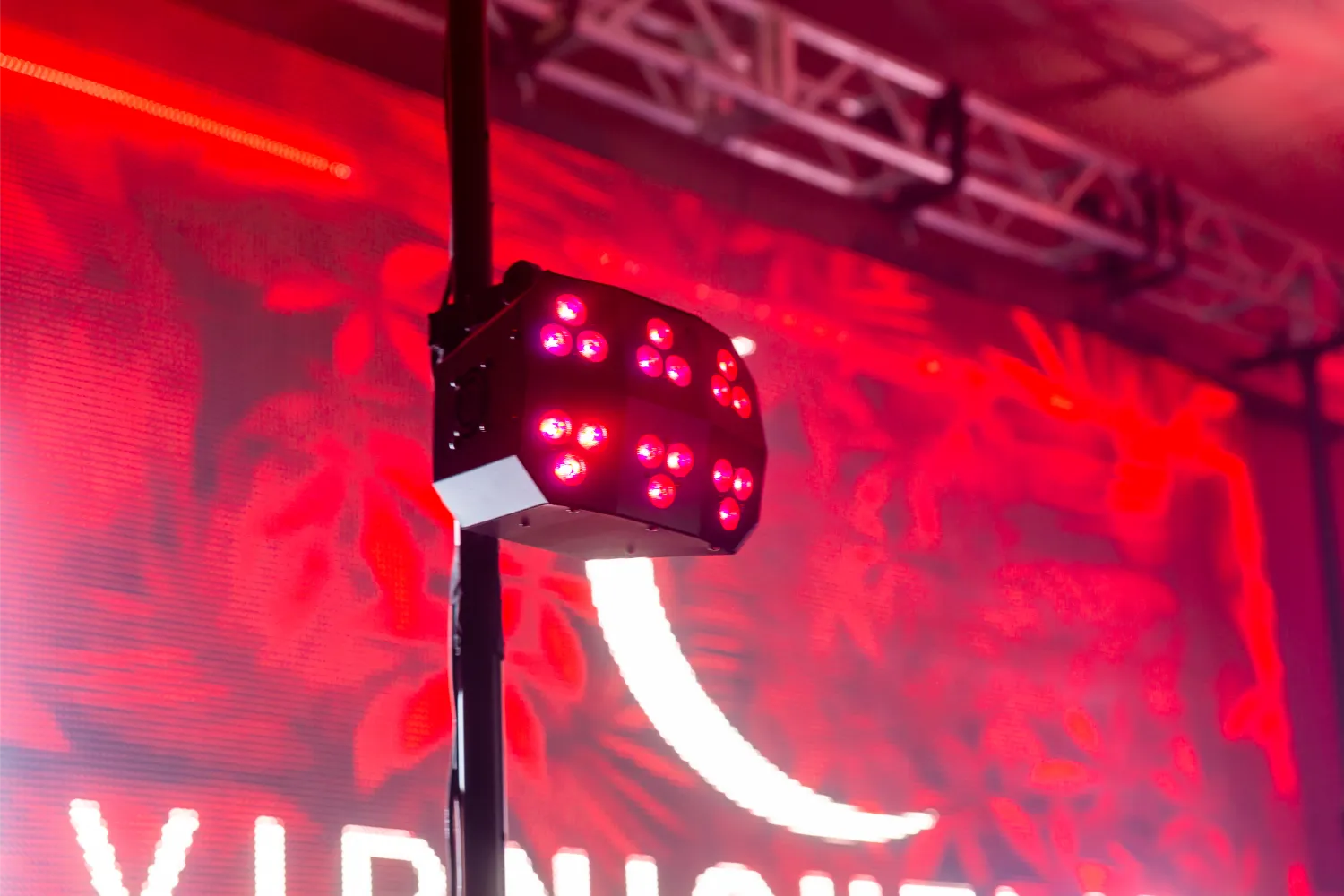 Black DJ wash light fixture with red LED beams mounted on a stand in front of a red illuminated background with partial text.