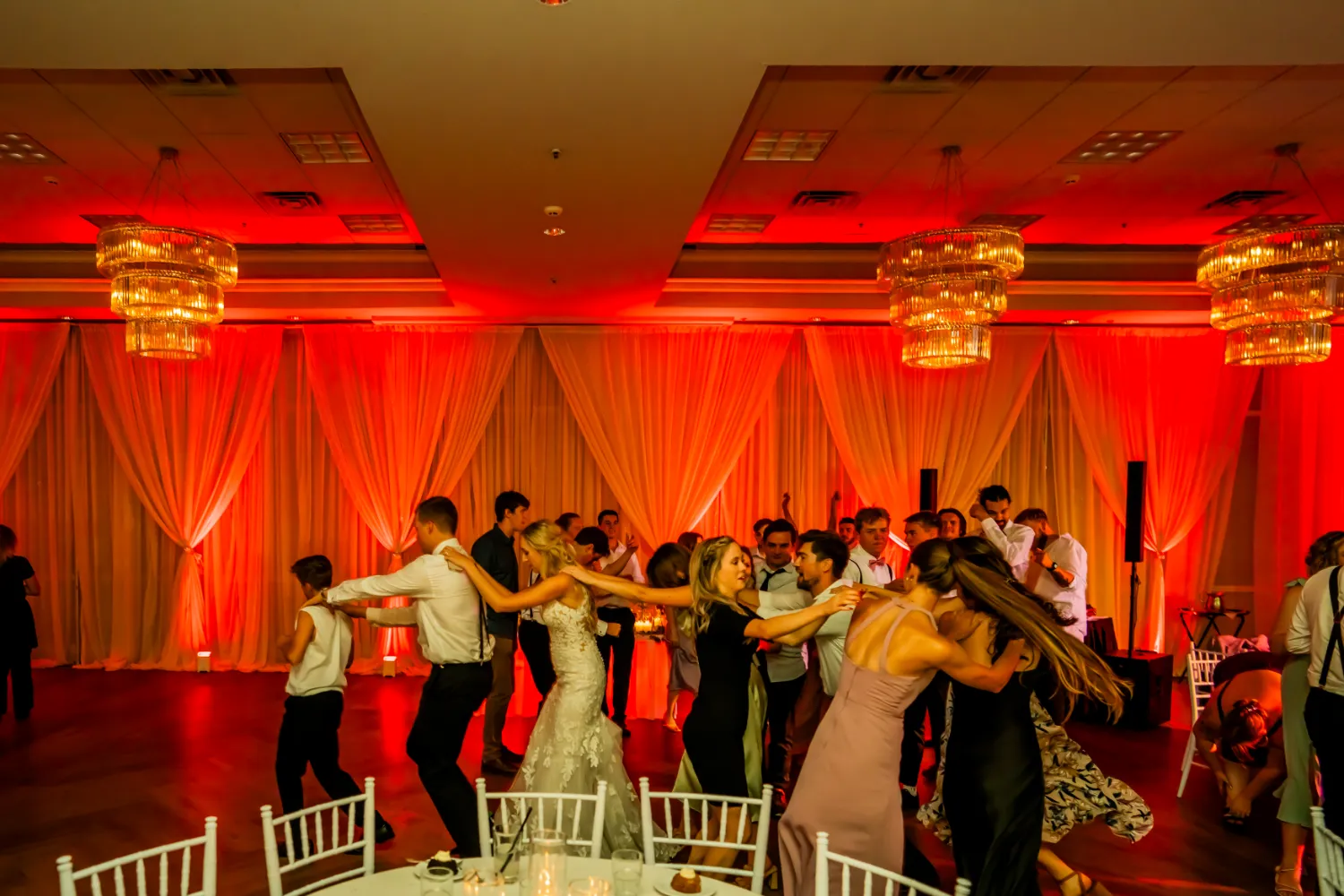 Guests dancing in a conga line at a wedding reception with warm red uplighting and chandeliers.