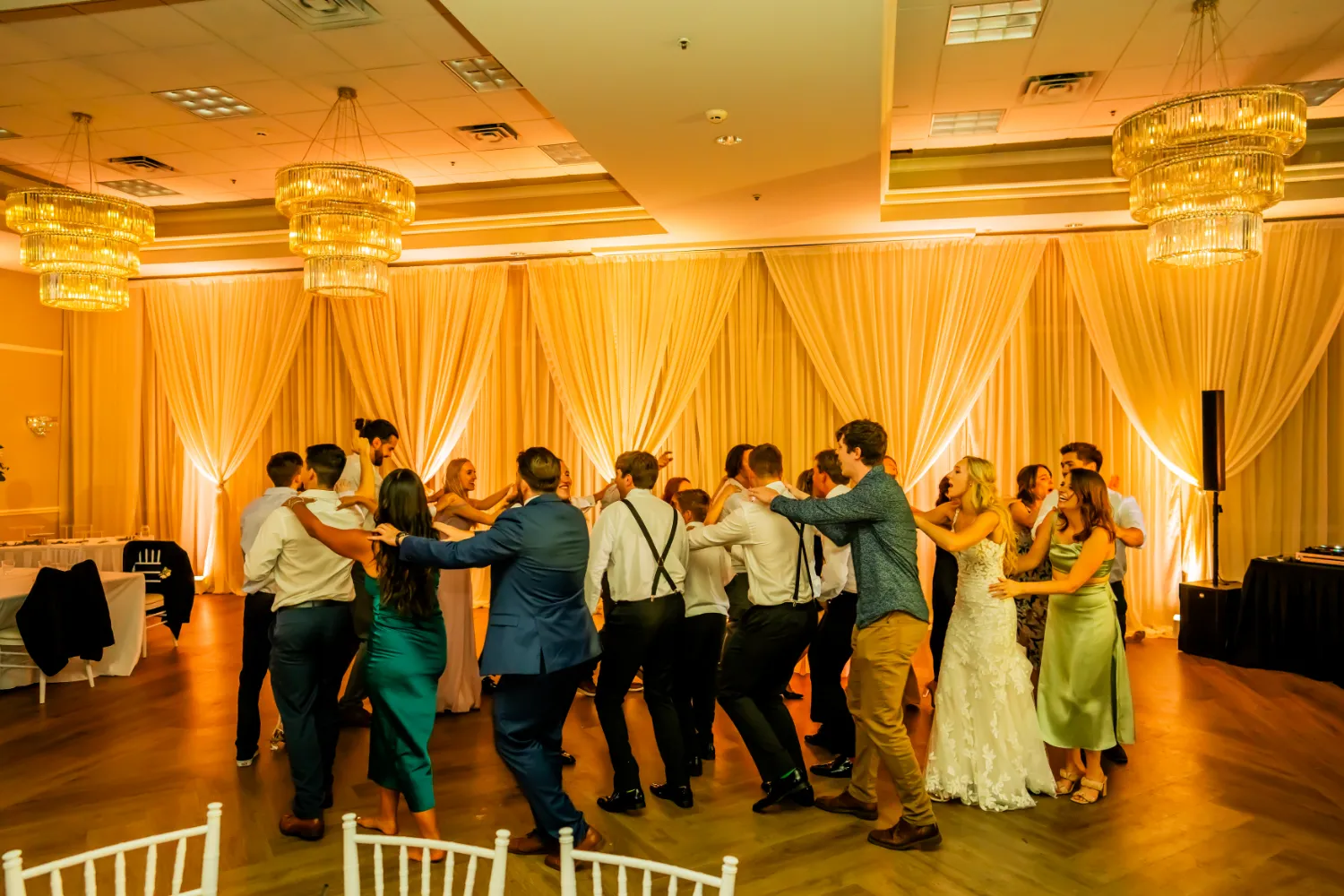 Group of wedding guests dancing in a conga line in a warmly lit banquet hall with chandeliers and draped curtains.