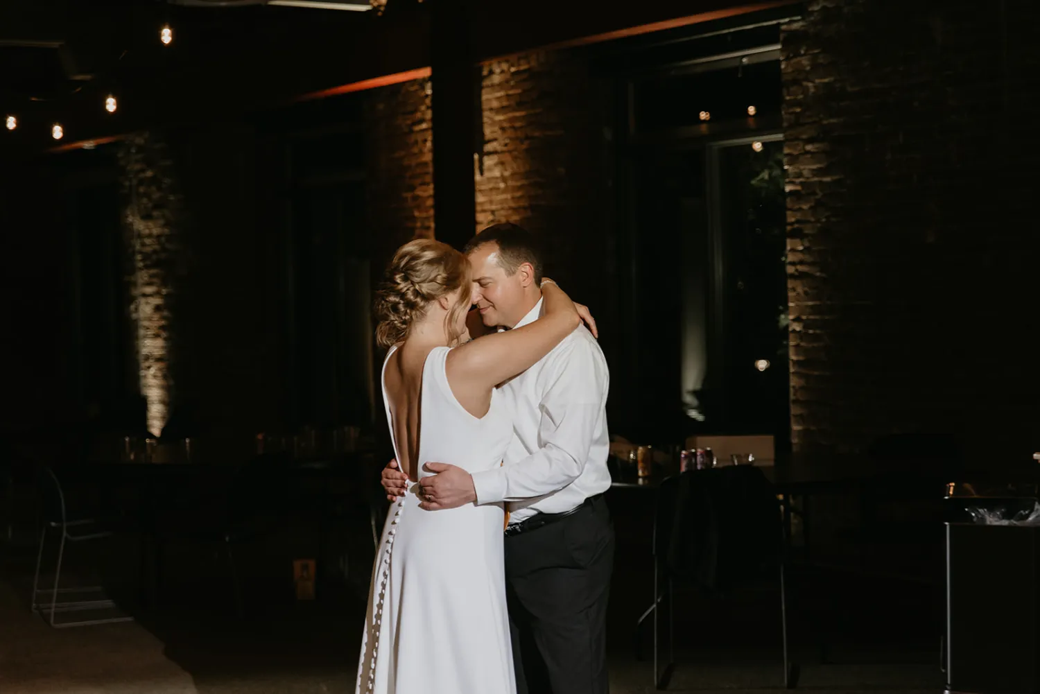 Bride and groom sharing an intimate first dance in a dimly lit venue with exposed brick walls.