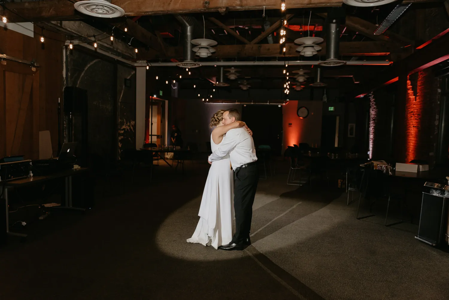 Bride and groom embrace while sharing a slow dance spotlighted in a dimly lit room with string lights and industrial decor.
