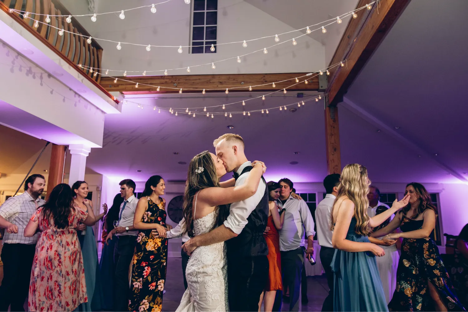 Bride and groom kissing while guests dance under string lights in a warmly lit wedding reception.