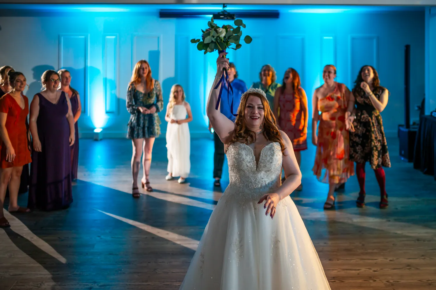 Bride in a white wedding dress holding a bouquet up, preparing to toss it to a group of women standing behind her in a dimly lit room with blue lighting.