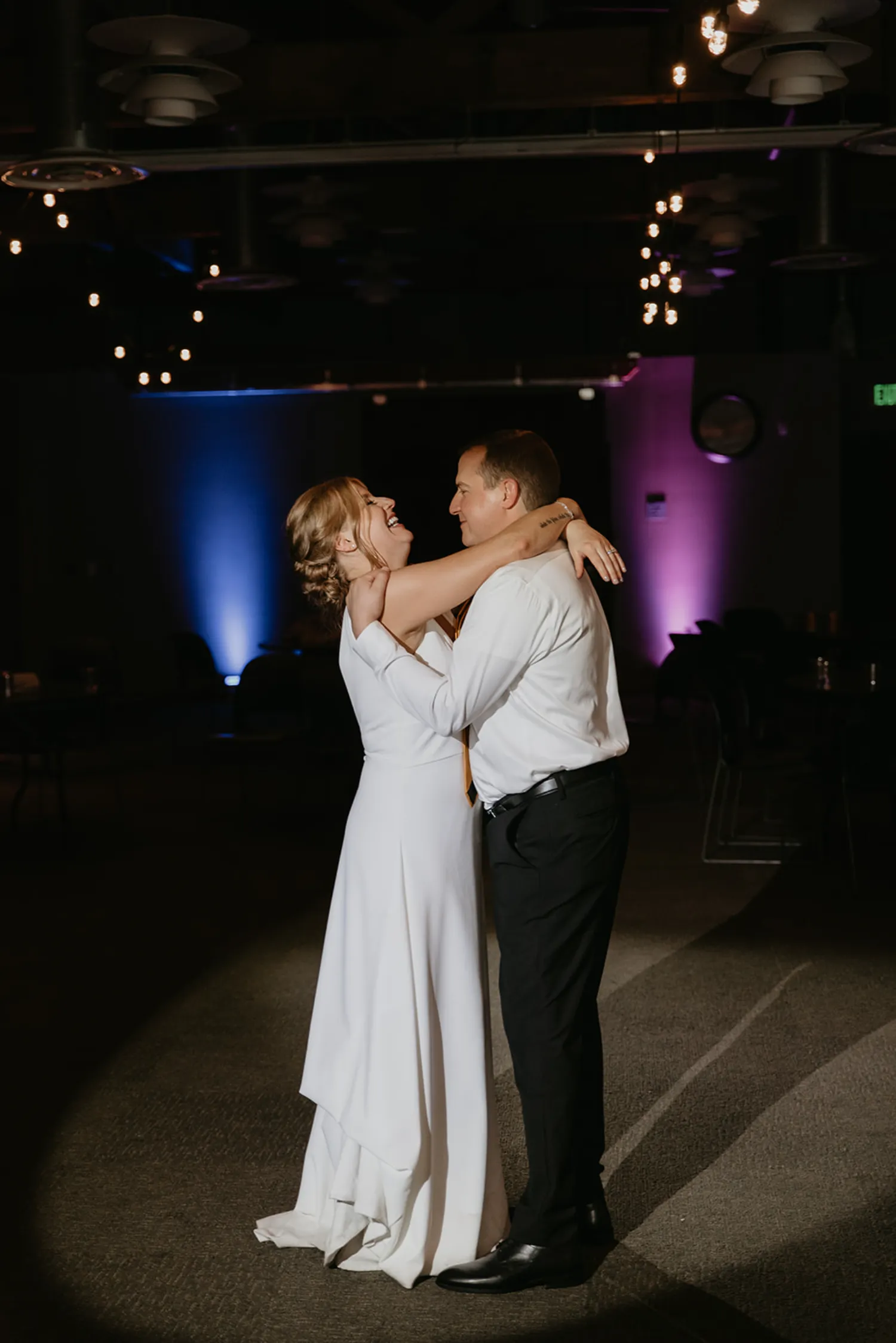 Bride and groom embracing and smiling during their wedding dance in a dimly lit room with decorative ceiling lights and blue and purple uplighting.