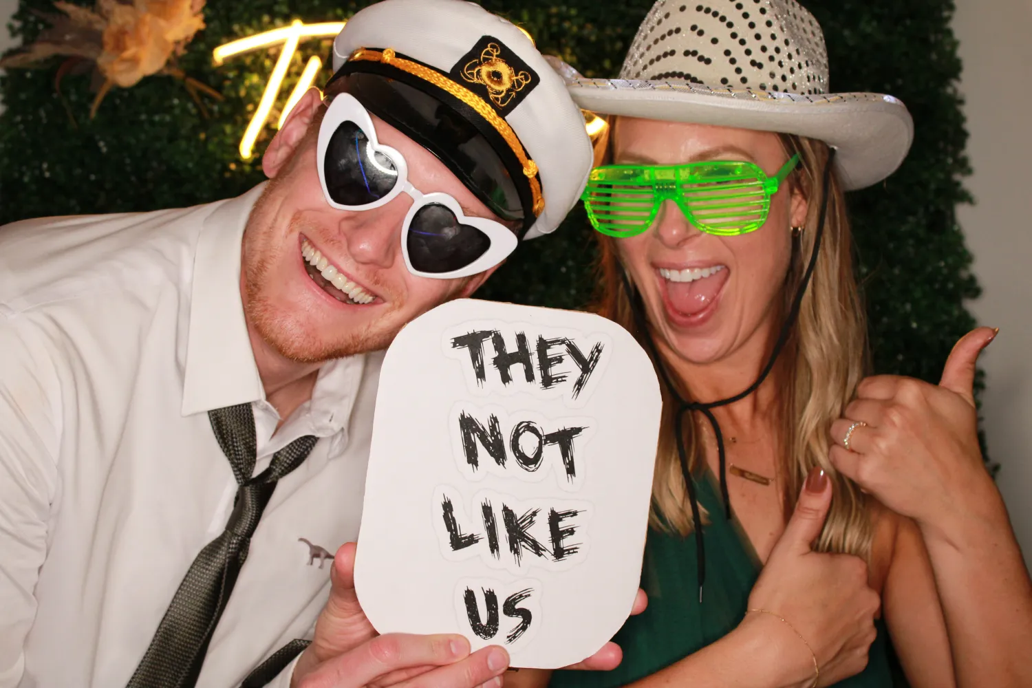 Two people in fun sunglasses and hats, holding "They Not Like Us" sign