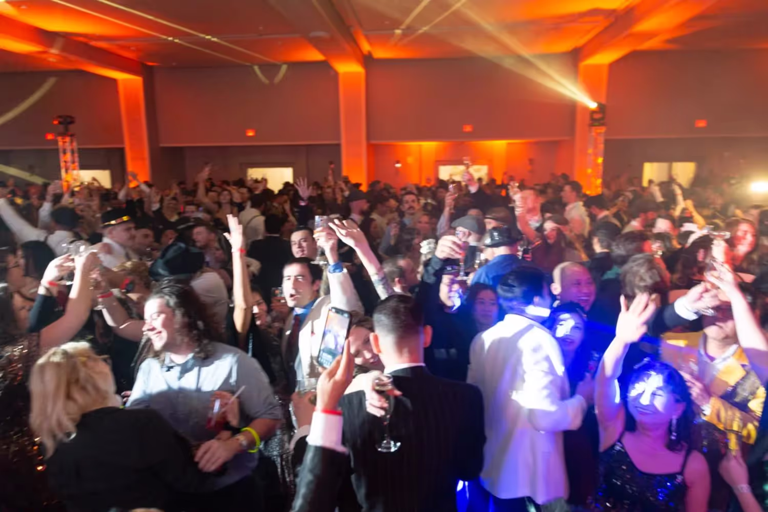 Crowded party with people dancing and raising drinks in orange-lit venue