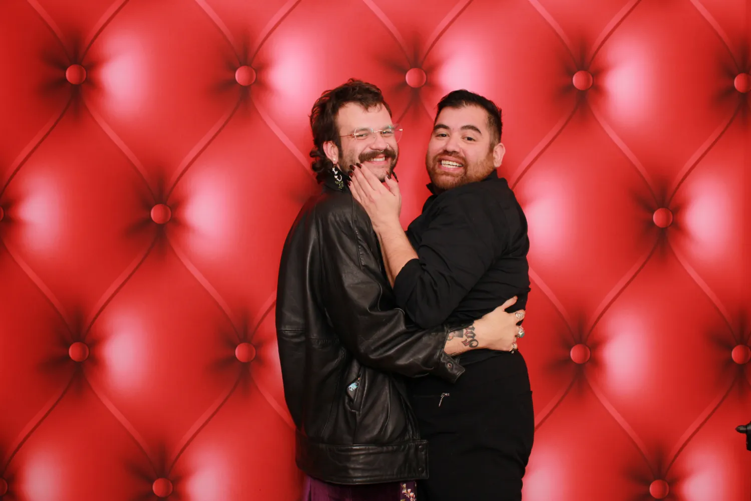Two people hugging and laughing in front of a red tufted background