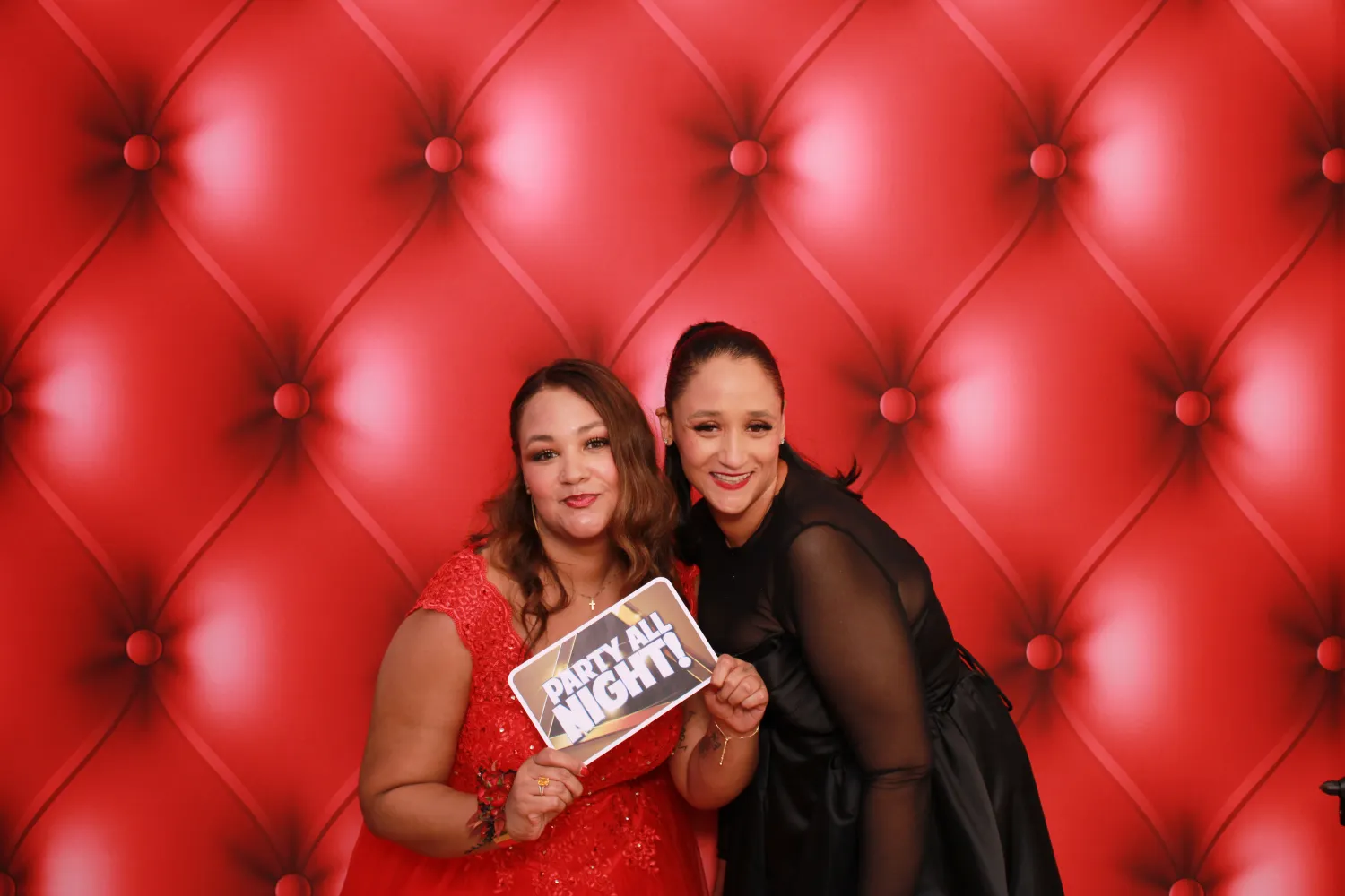 Two women posing with 'Party All Night' sign on red tufted background