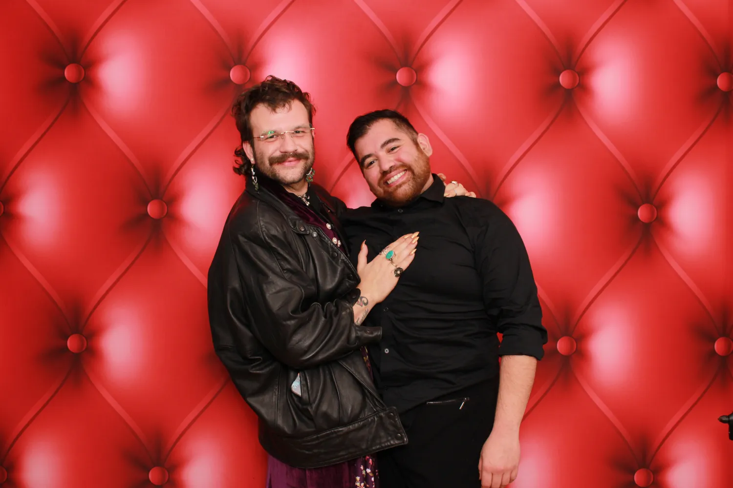 Two people posing together in front of a red tufted background