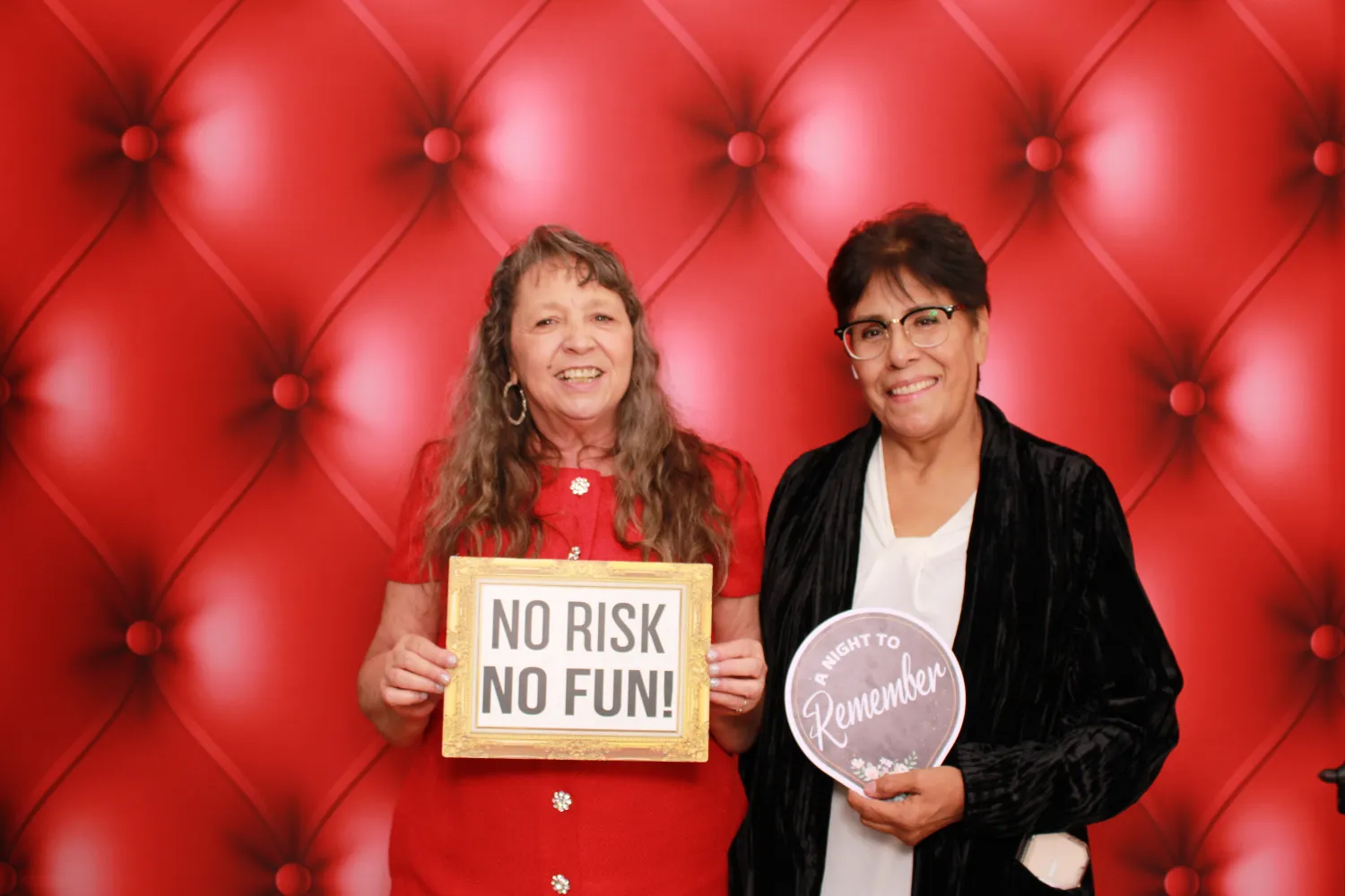 Two women pose with signs on red tufted background