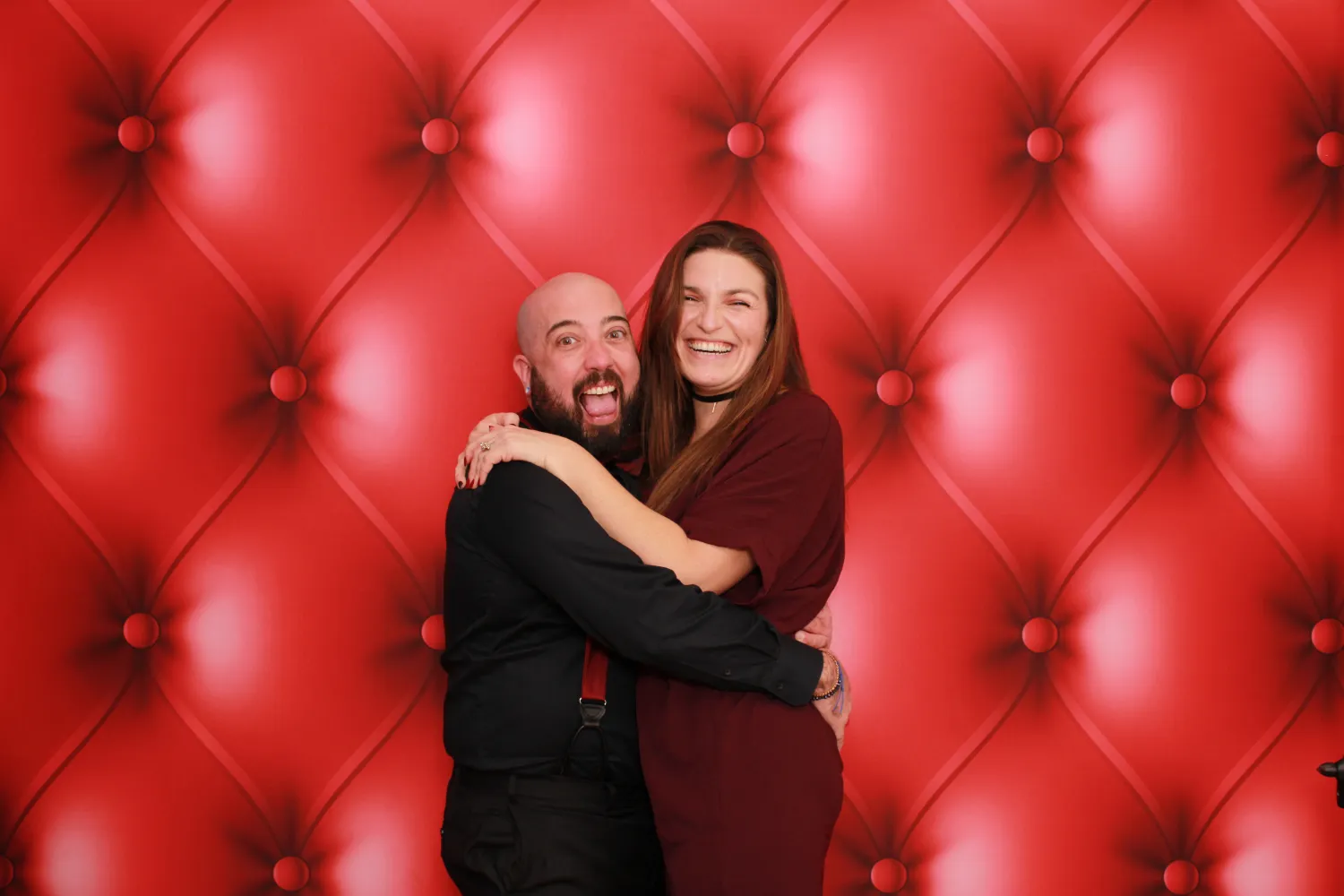 Two people hugging and laughing in front of a red tufted background