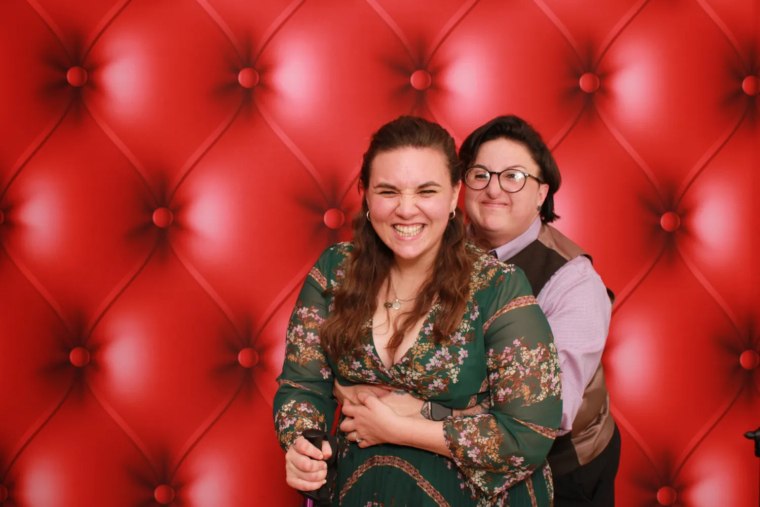 Two women smiling together in front of red tufted background