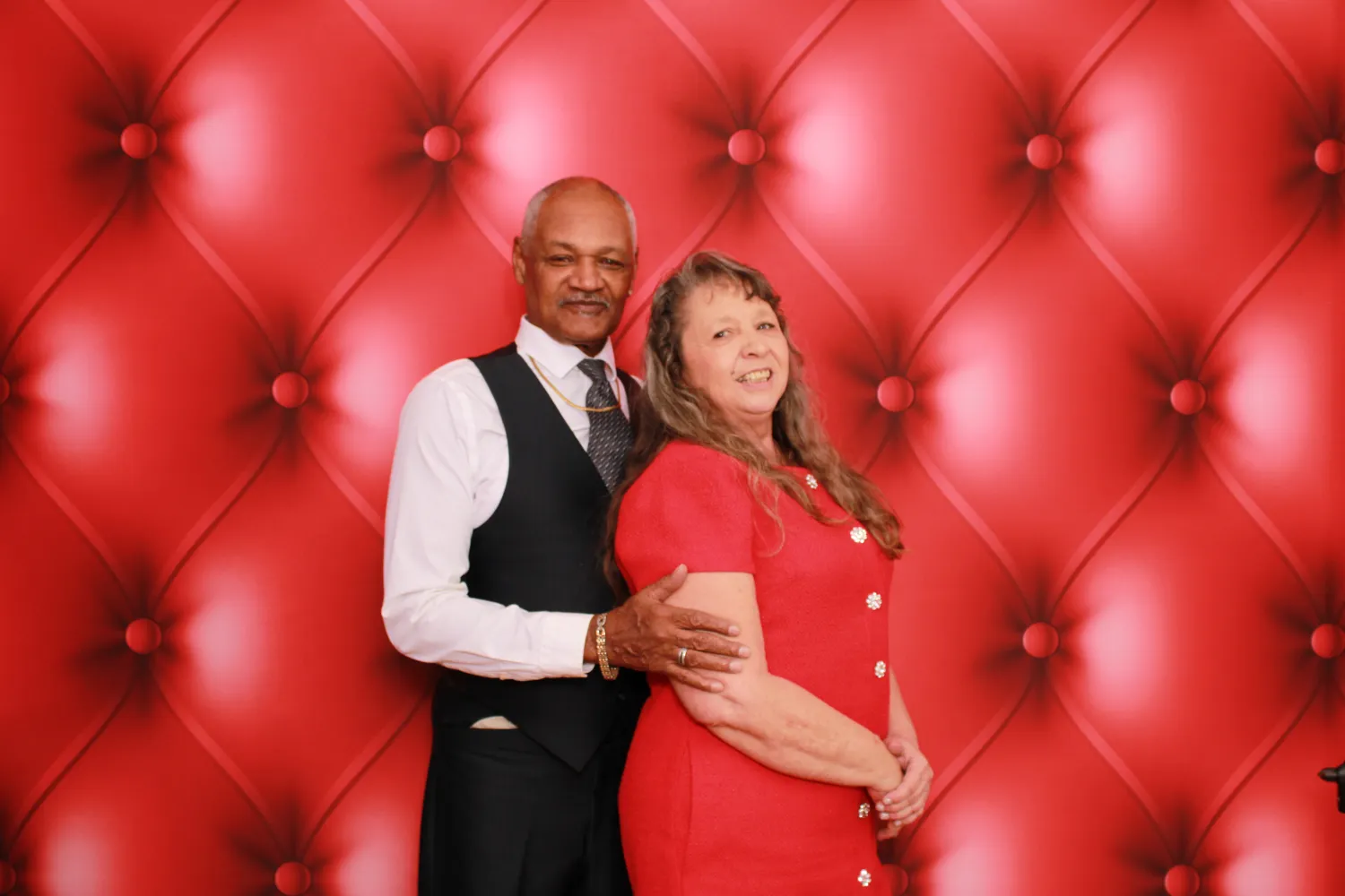 Couple in formal attire posing together against plush red tufted background