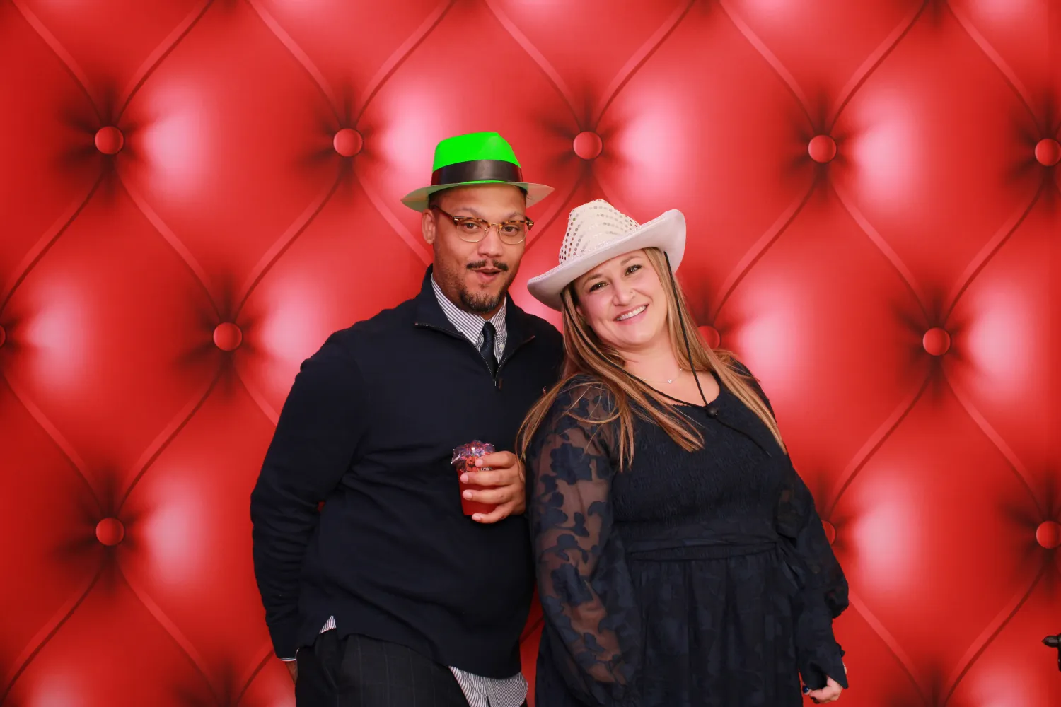 Two people in festive hats posing against a red tufted background