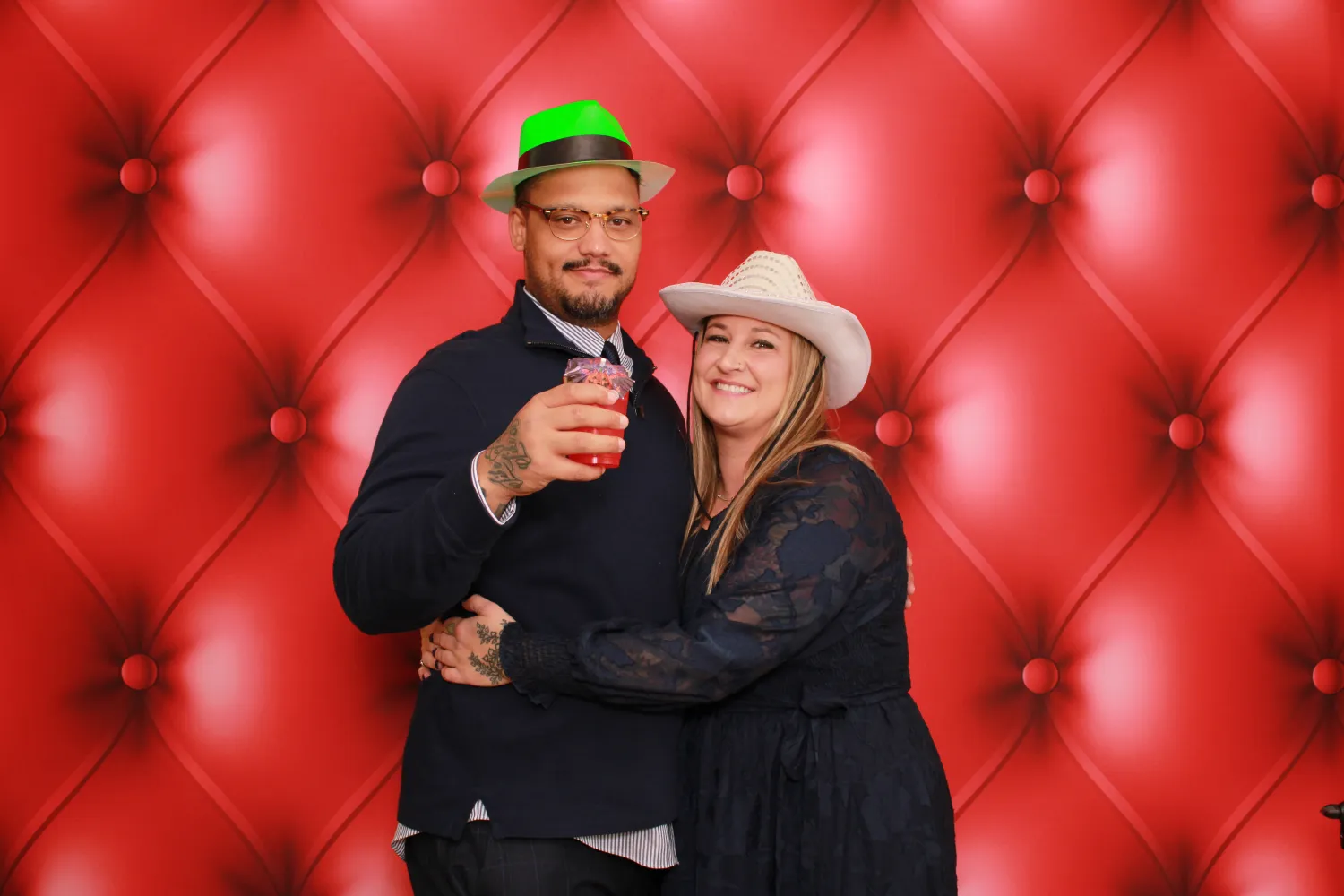 Couple in black outfits posing with drink against red tufted background