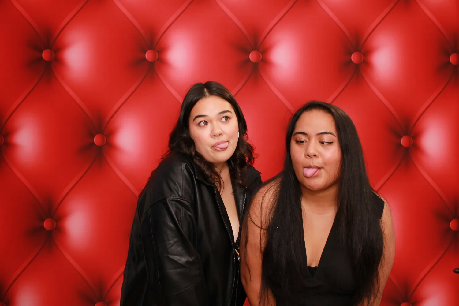 Two women posing with playful tongue-out expressions against red tufted background