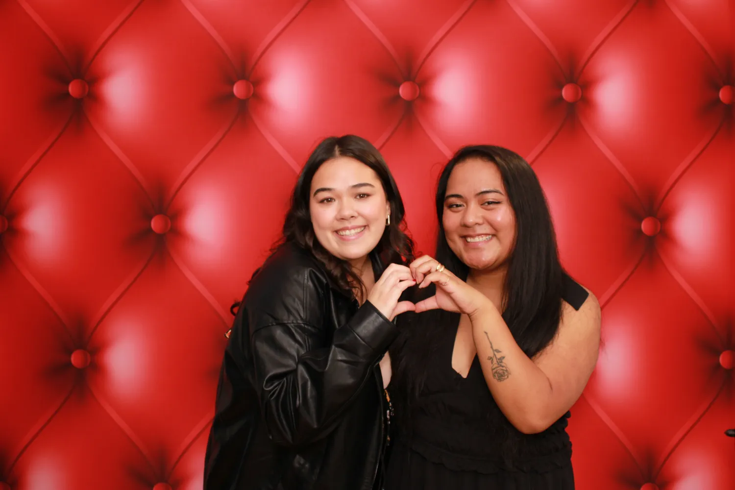 Two friends making heart shape in front of red tufted backdrop