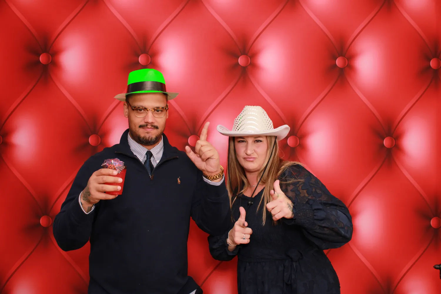Two people in different hats posing against red tufted background