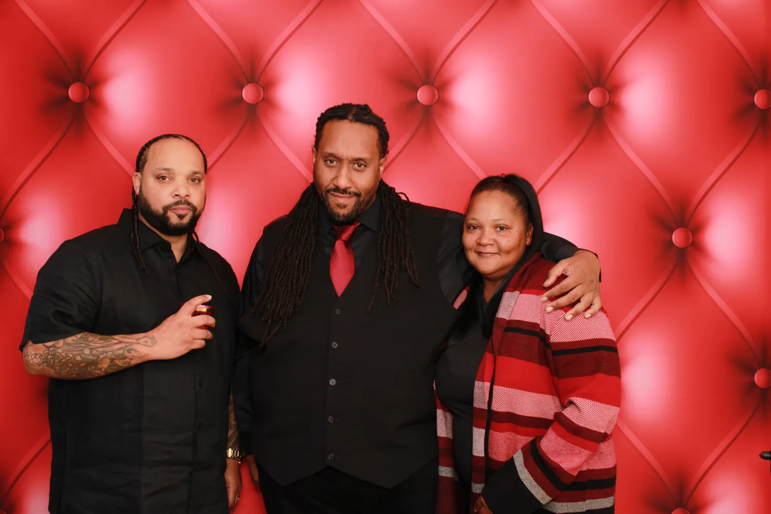 Three people posing against a red tufted background in black and red attire
