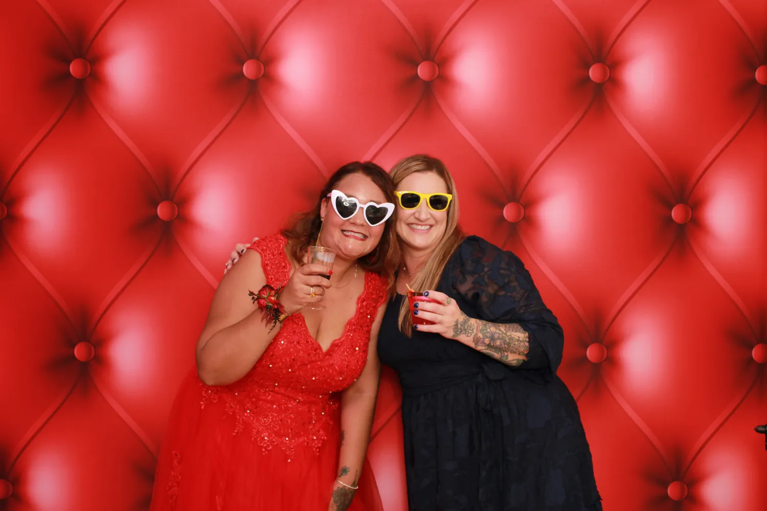 Two women in sunglasses posing together in front of red tufted backdrop
