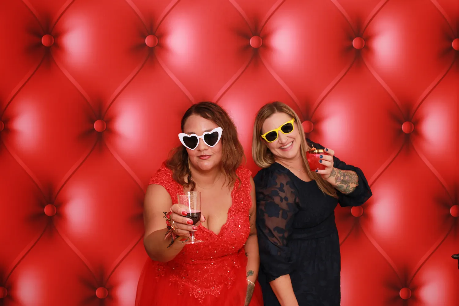 Two women in sunglasses posing with drinks against red tufted background