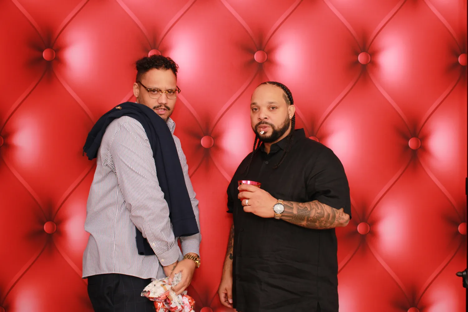 Two men pose in front of red tufted backdrop with holiday gifts
