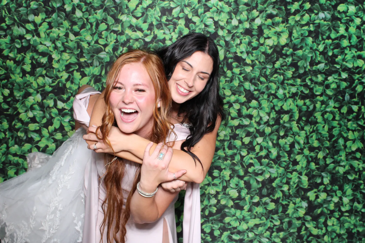 Two women laughing together in front of green leafy photo booth backdrop