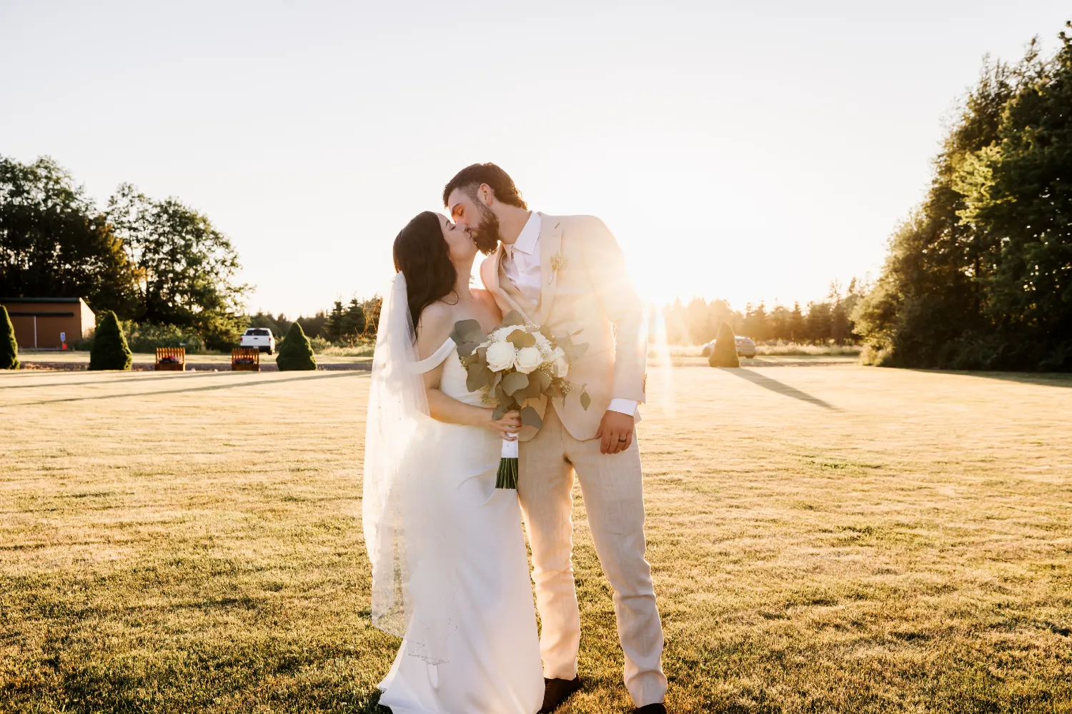 Newlyweds kissing at sunset on grassy field, bride holding white bouquet