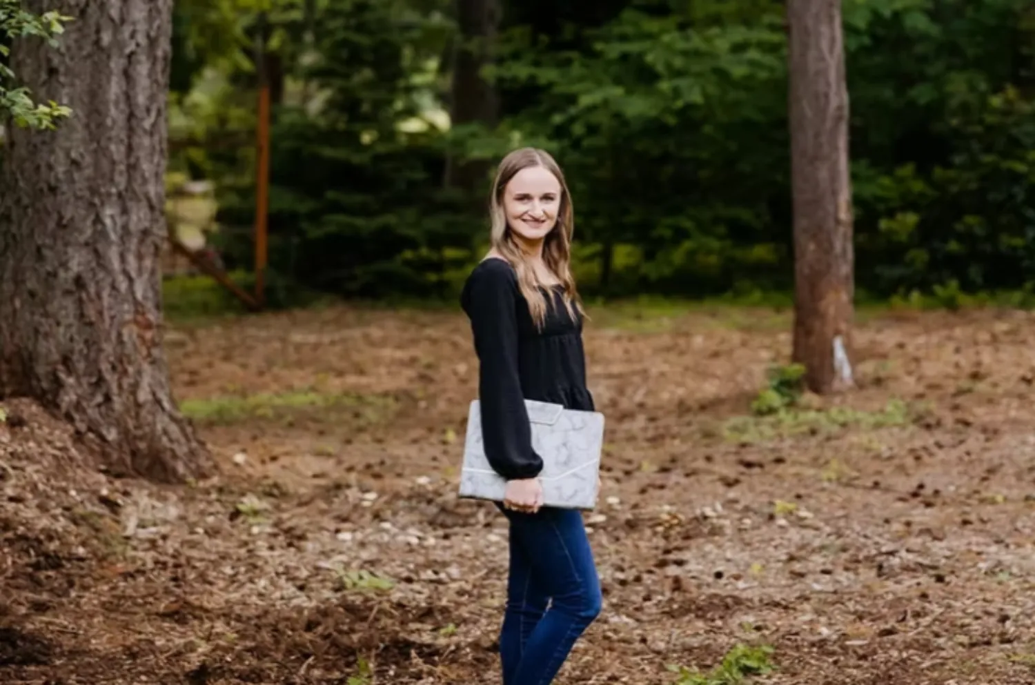 A person standing in a forest, holding a laptop, wearing black top