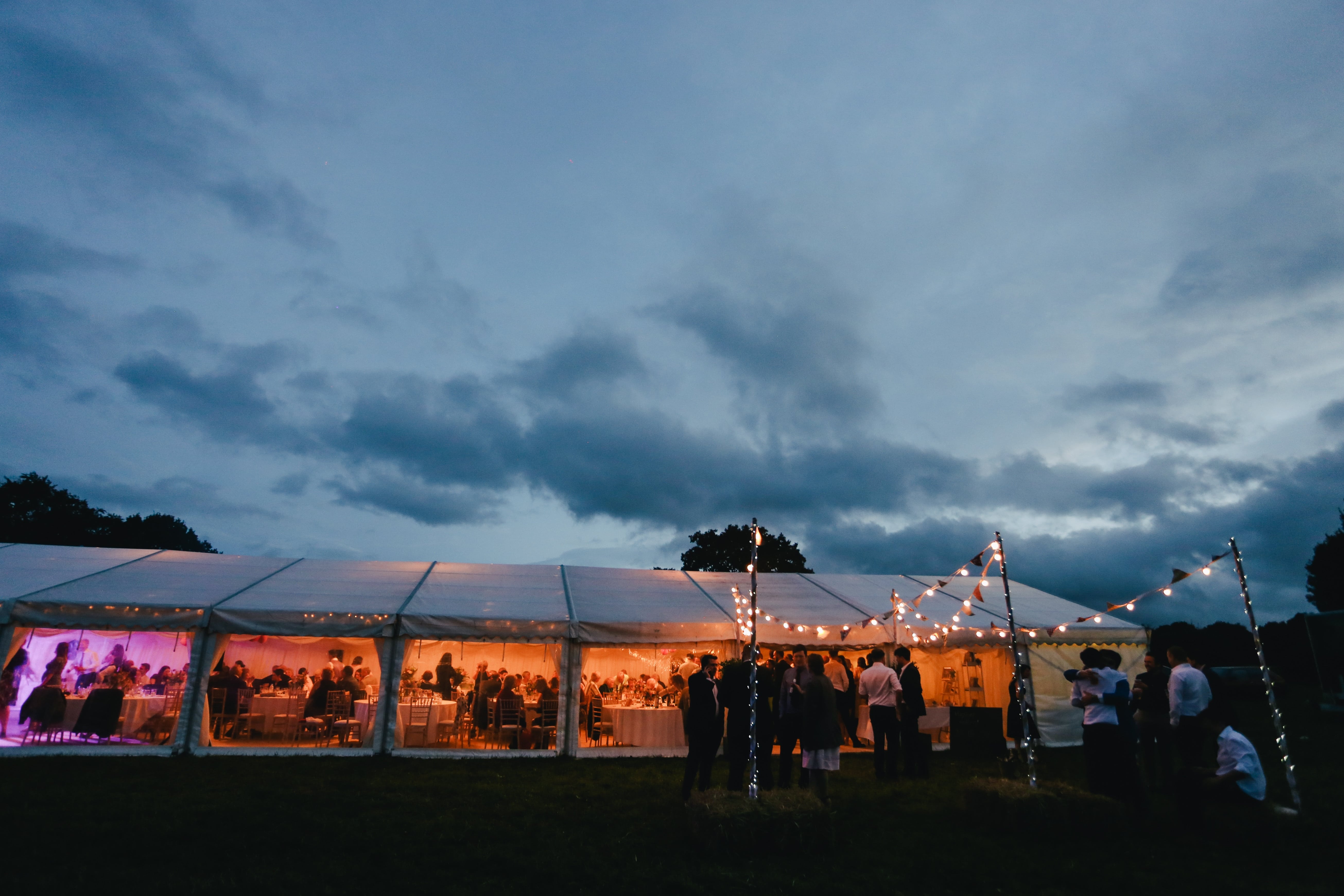 Evening wedding reception in a glowing tent with string lights under cloudy sky
