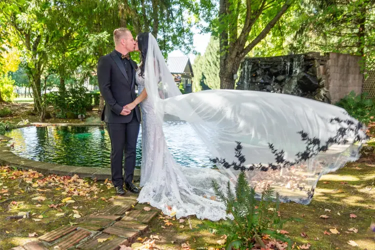 Bride and groom sharing a kiss beside the pond and waterfall at Water Oasis in Newberg, Oregon, with the bride's veil flowing gracefully in the breeze.