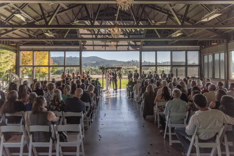 Bride and groom exchanging vows during an indoor ceremony at Water Oasis in Newberg, Oregon, with guests overlooking scenic vineyards and rolling hills through floor-to-ceiling windows.