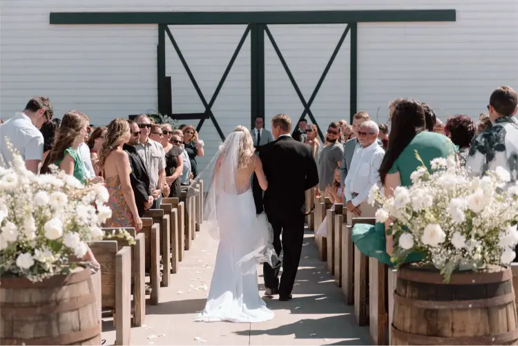 Bride walking down the aisle with her father during an outdoor Butler Barn wedding ceremony surrounded by guests and white floral arrangements.
