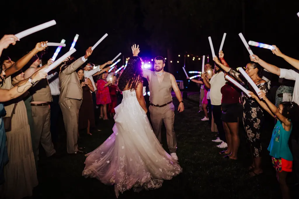 Bride and groom walking through a glow stick send-off surrounded by cheering guests during a fun nighttime wedding celebration at CedarVale Events.