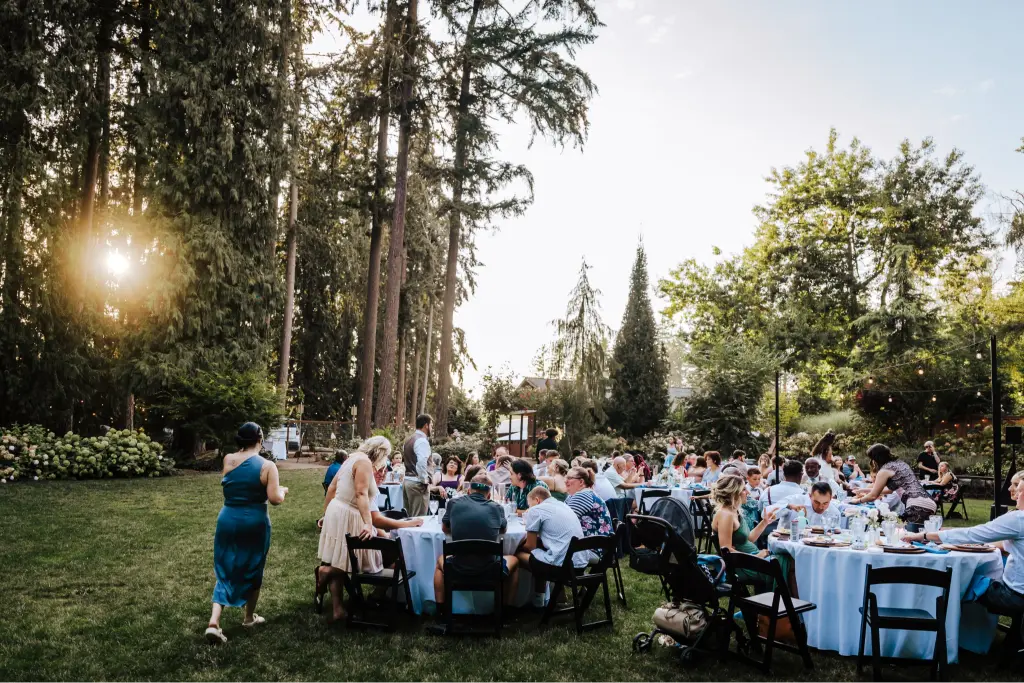 Guests enjoying an outdoor garden wedding reception at CedarVale Events with elegant tables, towering evergreen trees, and romantic string lights.