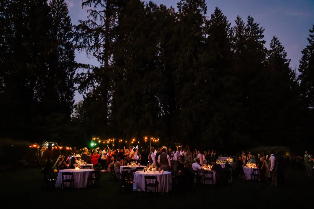 A wide view of an elegant evening wedding reception at CedarVale Events featuring candlelit tables, string lights, and guests celebrating beneath towering evergreen trees.