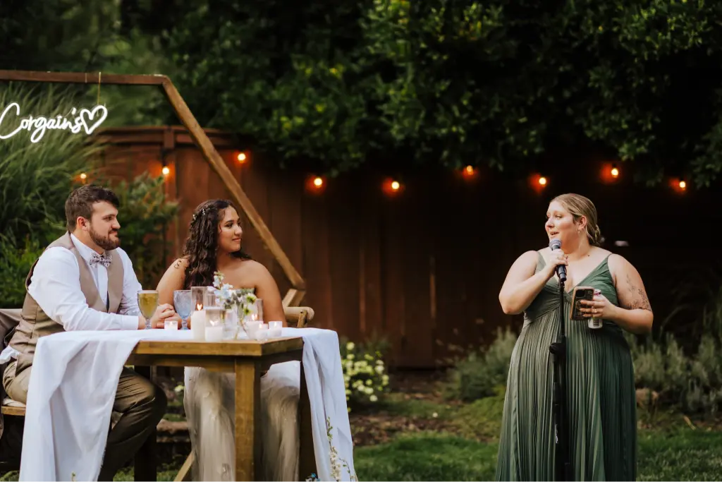A bridesmaid giving a heartfelt wedding toast while the bride and groom listen from their sweetheart table during an outdoor reception at CedarVale Events.