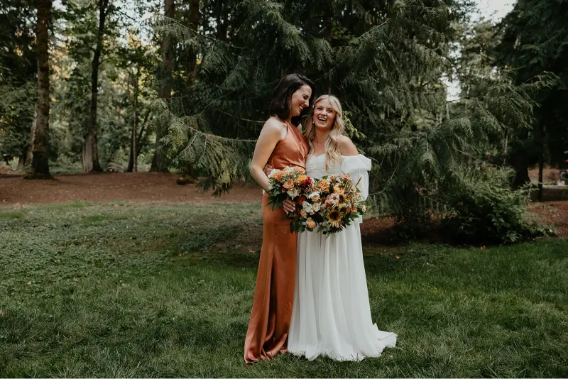 Bride and bridesmaid smiling together with colorful bouquets during a Wedding at Cedarville Lodge surrounded by lush forest scenery.